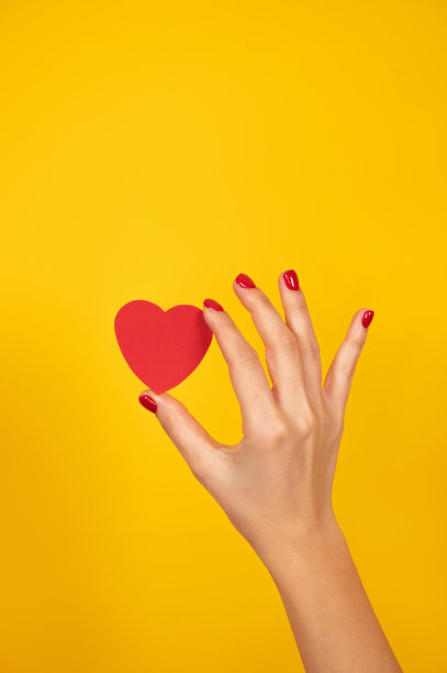 一个女人的手握着红色的指甲，在黄色的背景上拿着一颗纸心。爱情标志，情人节概念。图片下载