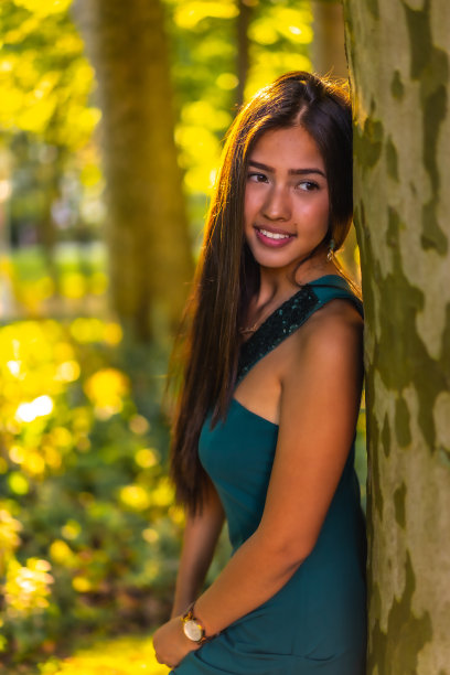 一个年轻的深色头发的拉丁人，长直发，穿着绿色的裙子倚在一棵树上。肖像沉思和感性的年轻女子，垂直照片图片下载