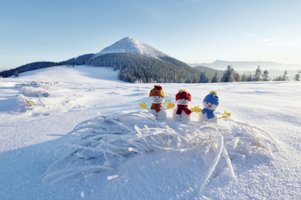 戴着帽子和围巾的小雪人在白雪覆盖的草坪上。美丽的冬季日落背景。有高山的景观。圣诞快乐，恭贺新禧。图片下载