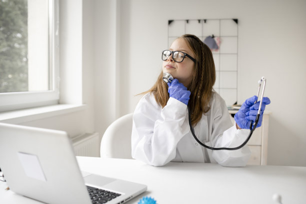 漂亮的金发女学生扮演医生，穿着白大褂，戴着蓝色防护手套，使用听诊器图片下载