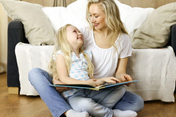 幸福的家庭。金发碧眼的年轻母亲坐在木地板上为她可爱的女儿读书。母亲的概念图片下载