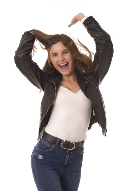 漂亮的女孩穿着皮夹克在白色的背景上玩和跳舞。穿着牛仔裤的女孩站在一个白色的演播室里，对着镜头摆姿势笑。图片下载