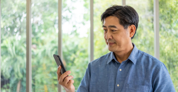 亚洲老人拿着智能手机模拟与空屏幕添加应用程序的标志，同时指着那个设备微笑。活跃的高级技术与模糊的绿色自然背景。图片下载