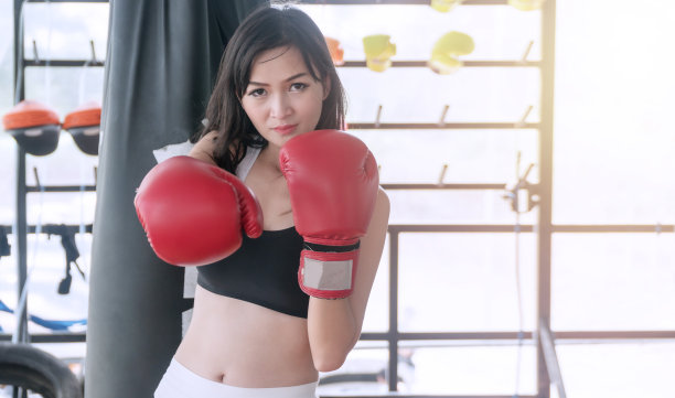 身材苗条的年轻女子穿着黑色的运动酒吧和红色拳击手套看着相机，而在体育健身中心的背景图片下载