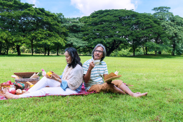 老年人生活方式的概念。幸福老两口背靠背坐在公园里一起放松。丈夫捧着一杯咖啡，妻子捧着一把刀，手里拿着橘子皮，在草地上野餐。图片下载