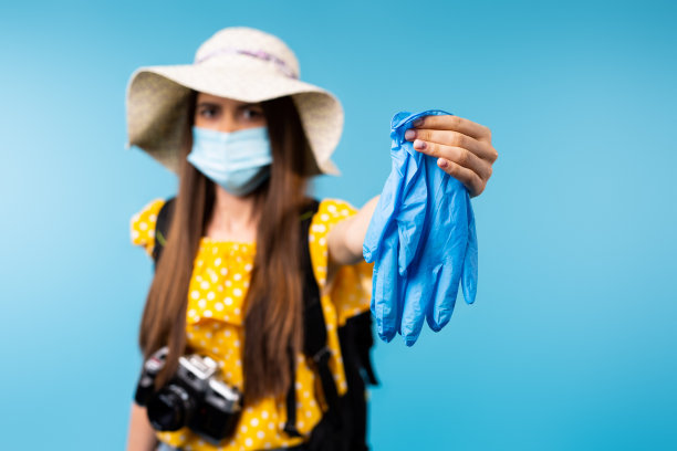 屏幕上是一张模糊的照片，一个戴着帽子和医用口罩的女游客伸出乳胶防护手套。蓝色背景。大流行和旅行。图片下载