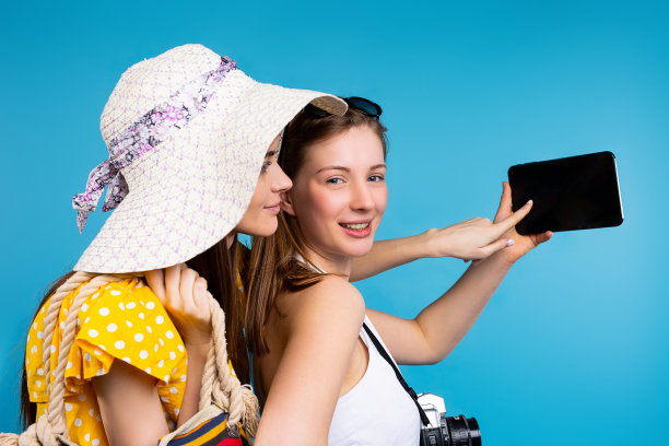 照片中，蓝色背景下的两位女友穿着夏装，手拿平板电脑，食指指着屏幕。图片下载