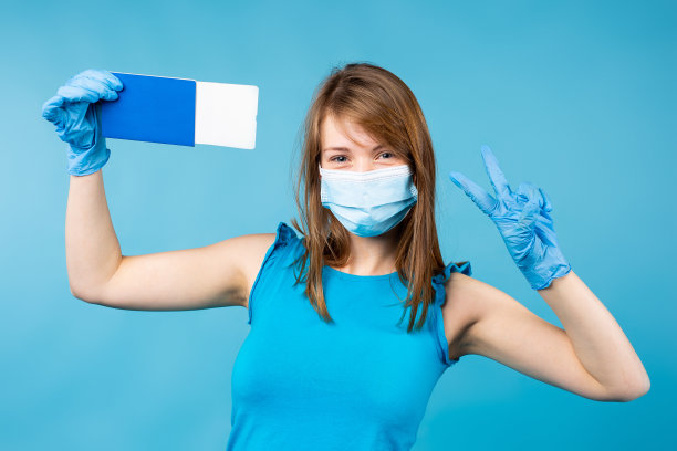 旅游大流行。假期安全旅行。黑白照片的漂亮女人穿着蓝色衬衫，保护蓝色面具，手套。拿着蓝色护照和机票，在蓝色上做着和平的手势图片下载