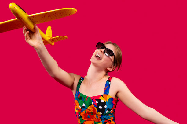 一个快乐的女孩穿着鲜艳的衣服，在阳光的手里拿着一架黄色的飞机在红色的背景和梦想的旅行。旅游与旅游广告空间。图片下载