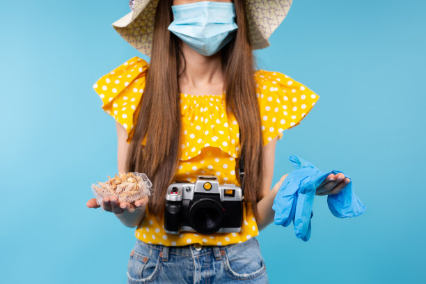 裁剪照片的黑发妇女与照片相机戴着医疗防护口罩和夏季帽子，拿着蓝色医疗手套和贝壳。蓝色背景下的流感假期图片下载