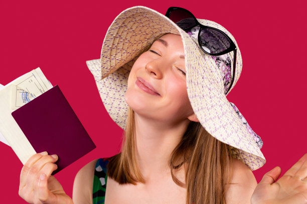 一个心满意足的白人女孩戴着一顶帽子，闭着眼睛，手里拿着护照和旅行用的国际疫苗接种证书。红色的背景。图片下载