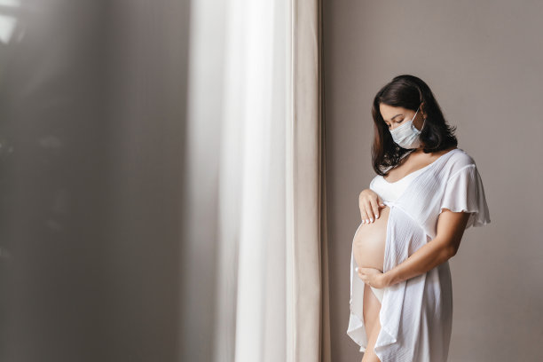 年轻美丽的孕妇戴着医用外科口罩在家靠窗。图片下载