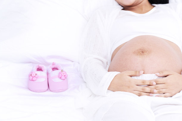 年轻孕妇躺在白色的床上，穿着粉红色的婴儿鞋。在卧室里用童鞋封闭腹部的人们。图片下载