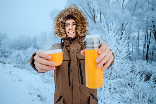 一个穿着带帽羽绒服的快乐男人在寒冷的暴风雪和霜冻过后，在结霜的树木背景下，从保温瓶里喝着热茶图片下载