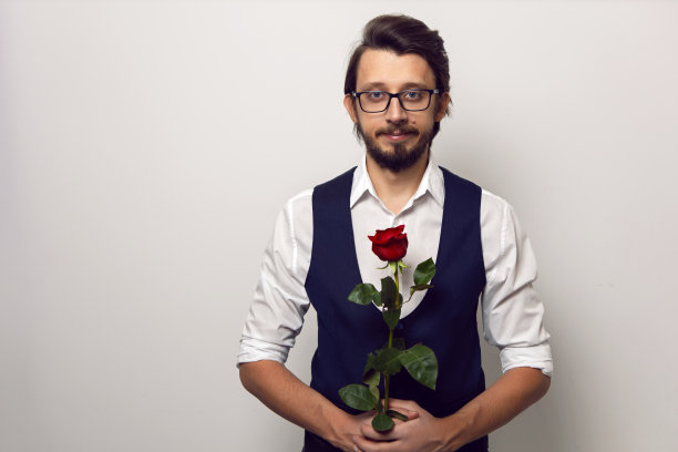 情人节那天，一名商人戴着眼镜，留着胡子，身穿白色衬衫和背心，白色背景下站着一朵红玫瑰图片下载