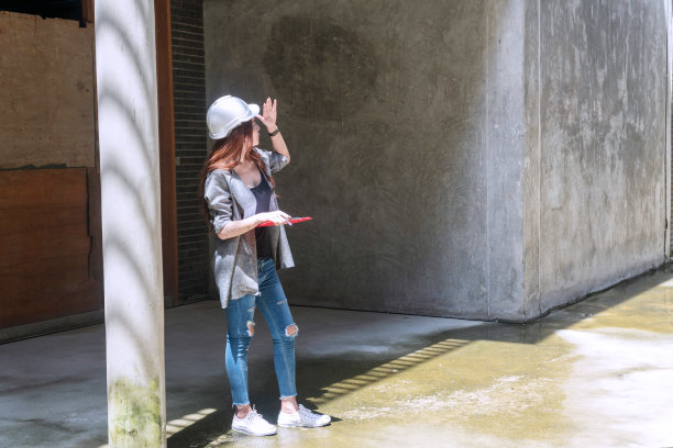 工程师年轻女子戴着白色头盔站在户外，手持文件夹文件现场工作。女建筑师戴着施工安全帽或安全帽在工地检查正在进行的工作。图片下载