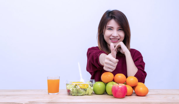 健康食品的概念。快乐的年轻女子早餐与沙拉素食，水果和果汁在白色的背景。微笑的女孩吃着新鲜的健康沙拉，苹果和橘子竖起大拇指。图片下载