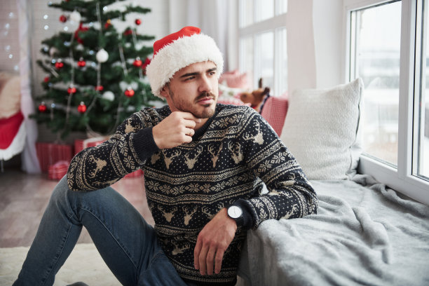 一个戴着圣诞帽、穿着节日服装的男人透过窗户往外看。背景上的圣诞树图片下载