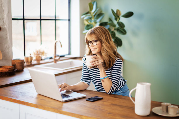 这张照片拍摄的是一位中年妇女在厨房边喝咖啡边用笔记本电脑工作。家庭办公室。图片下载