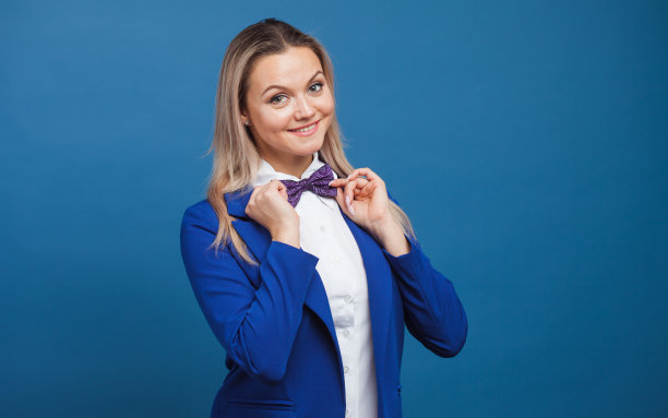 蓝色背景上穿着蓝色衣服的友好的年轻女人。穿夹克打着领结的金发女孩图片下载