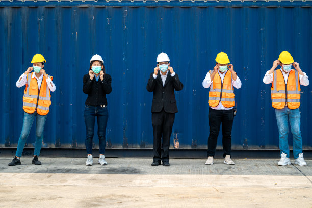 群人工作人员戴着防护面具、安全帽，穿着工作服、安全服，背景是货仓。产业工人操作。人们有一个社交距离空间。图片下载