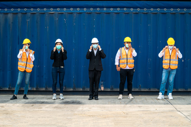 群人工作人员戴着防护面具、安全帽，穿着工作服、安全服，背景是货仓。产业工人操作。人们有一个社交距离空间。图片下载
