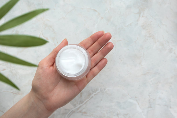 女士手持保湿霜在豪华大理石背景与复制空间，散焦热带树叶。卫生产品。护肤美容产品。保湿剂的概念。卫生保健的概念。图片下载