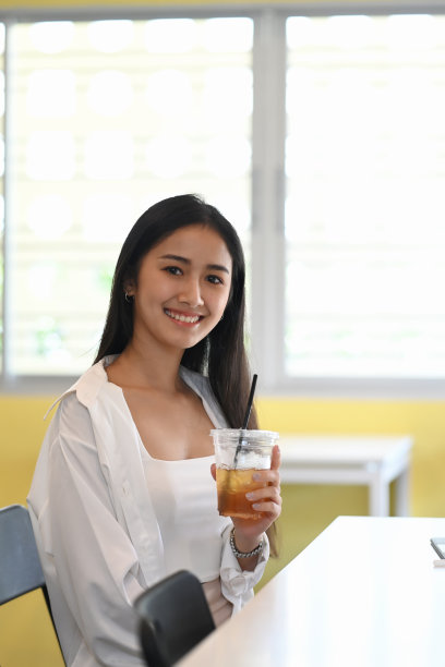 一个愉快的年轻女子坐在办公室里拿着冰咖啡微笑着对着镜头的肖像。图片下载