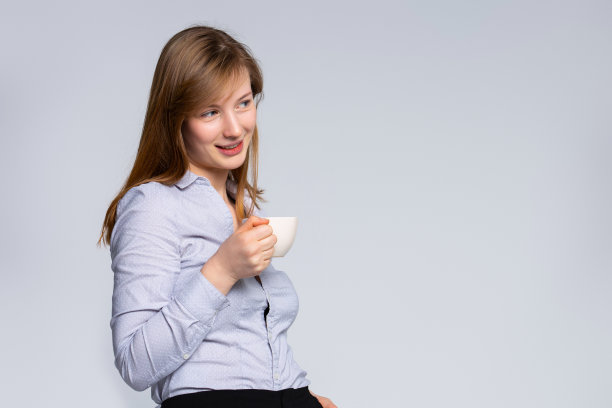 女商人拿着咖啡杯站着，看着远处。灰色背景上的空白。公司领导在工作中休息，幻想成功，考虑投资决策。图片下载