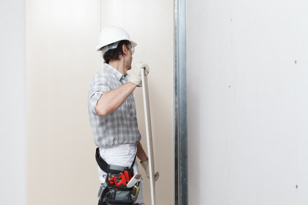 在墙上安装石膏板的工人。戴安全帽、工作手套、安全眼镜和工具带。孤立在白色背景与复制空间图片下载