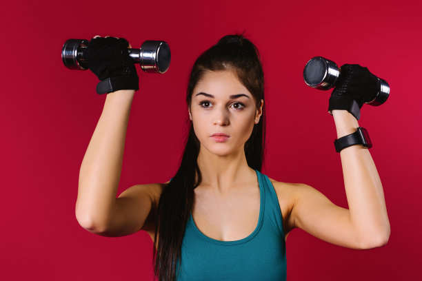 白人运动女孩在一个红色的背景上穿着上衣和铁哑铃摇动她的手臂肌肉。运动的概念和美丽的膨胀的身体。图片下载