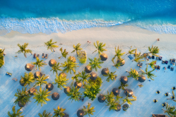 日落时分，印度洋沙滩上的雨伞、棕榈树鸟瞰图。夏天在非洲的桑给巴尔。热带景观有棕榈树，阳伞，行走的人，蓝色的水，波浪。俯视图图片下载