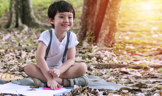 兴高采烈的小男孩拿着橡皮泥看着相机坐在公园里。小混合种族学校的孩子发射在他的手橡皮泥，而玩在自然的背景。图片下载