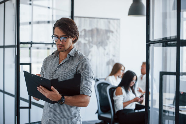 一个戴眼镜，手里拿着笔和笔记本的人站在对面的办公室里图片下载