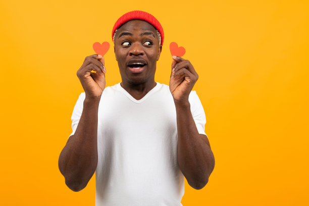 一张英俊的美国人的肖像，皮肤黝黑，身穿白色t恤，手里拿着两张小小的情人节心形明信片，看着黄色背景上的一边图片下载