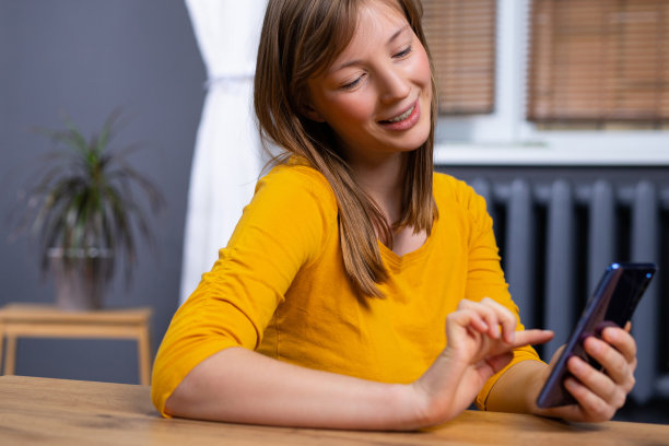 年轻美丽的女人兴奋的照片使用智能手机，而坐在室内窗户附近。在线学习的概念图片下载