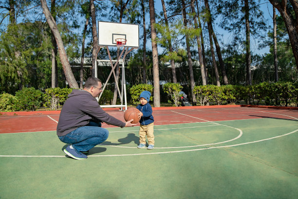 在树和蓝天的背景下，一个男人坐在操场上给一个小男孩打篮球图片下载