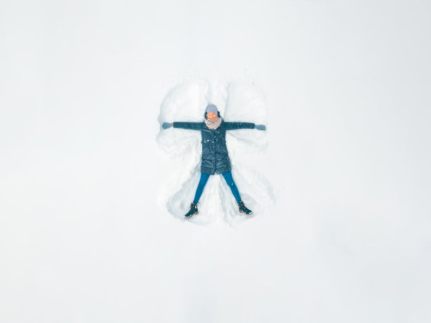 年轻漂亮的女人穿着灰色夹克，帽子和手套，蓝色牛仔裤，靴子在雪地里做一个雪天使，从无人机上看。雪娱乐,娱乐图片下载