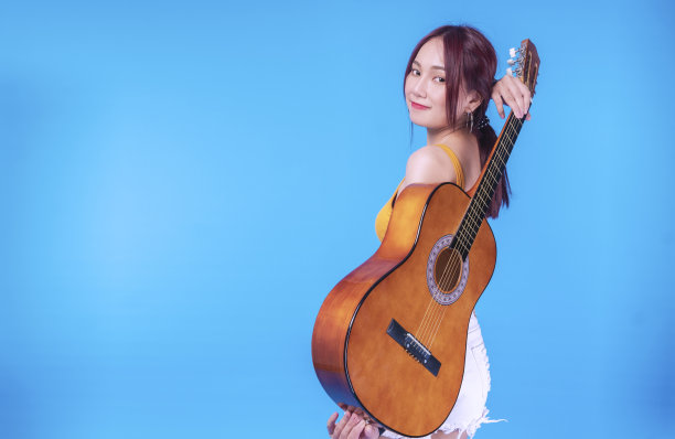 一个快乐的年轻女子，在她背后的原声吉他在蓝色的背景。肖像欢快的女性拿着吉他在她的背后，而站在孤立的背景。图片下载