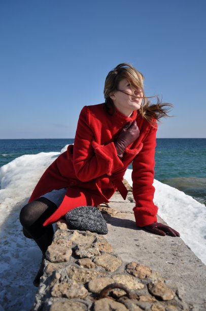在刮风的天气里，穿着红色外套、留着长发的年轻女子坐在海边的石头上图片下载