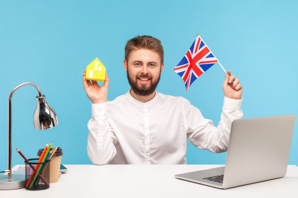 一个自信的蓄着胡须的男人坐在办公室里，拿着笔记本电脑，举着英国国旗和纸房子，梦想着在英国买个住处图片下载