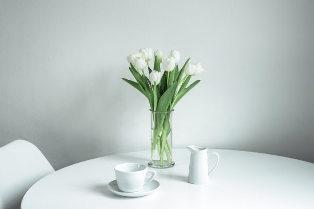 一束白色郁金香放在一个玻璃花瓶，一个杯子和一个牛奶罐在白色的桌子上图片下载