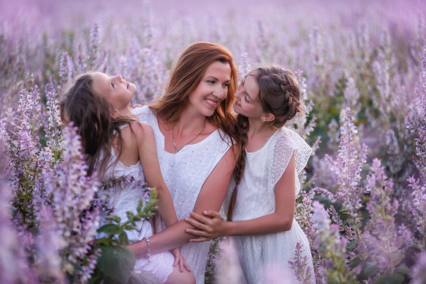 年轻的母亲抱着两个女儿在紫色盛开的鼠尾草田。女孩们在一起拥抱，欢笑，玩得开心。身穿白衣，头戴草帽。带着孩子旅行，关爱母亲。特写肖像图片下载