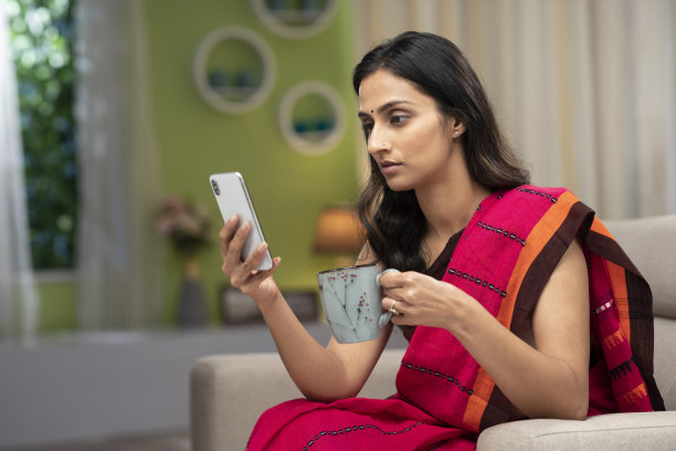 一个年轻漂亮的女人用手机和喝咖啡，坐在家里的沙发上，库存照片图片下载