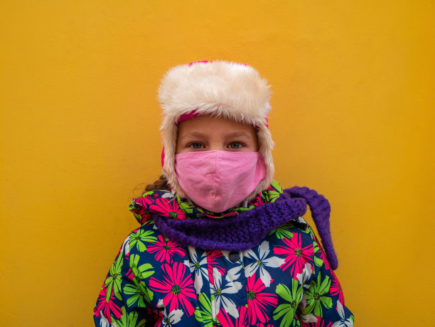 年轻的女孩在温暖的冬天的衣服与保护粉色面膜在黄色的墙壁背景。城市风格街景照片。图片下载