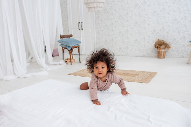 在极简主义的室内设计中，一个卷头发的非洲裔美国女孩站在一张白色的床旁。明亮通风的房间，斯堪的纳维亚风格。宝宝在家里玩耍，玩得开心。生活方式图片下载