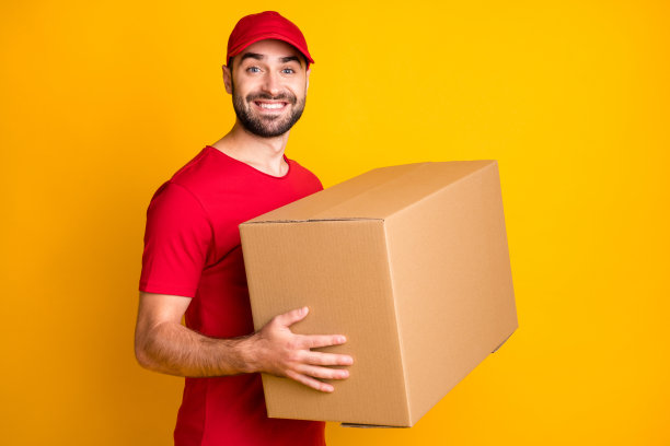 照片可爱的快递员穿着红色的衣服微笑着拿着一个大的棕色盒子孤立的黄色背景图片下载
