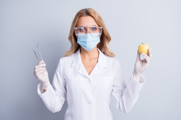 照片的成熟的牙科妇女戴着口罩和白色外套持有口腔医学设备和苹果孤立在灰色背景图片下载