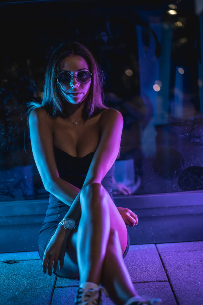 一位身穿黑色连衣裙的年轻白人女性，在紫色和蓝色LED灯反射的黑色水晶的照射下，静静地坐在地板上。夜晚城市摄影图片下载