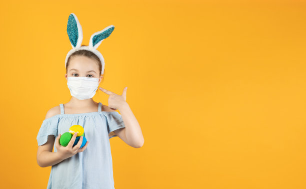 一名女孩戴着冠状病毒的医用口罩，头上戴着兔耳，手里拿着不同颜色的复活节彩蛋。复活节和春天的象征。图片下载